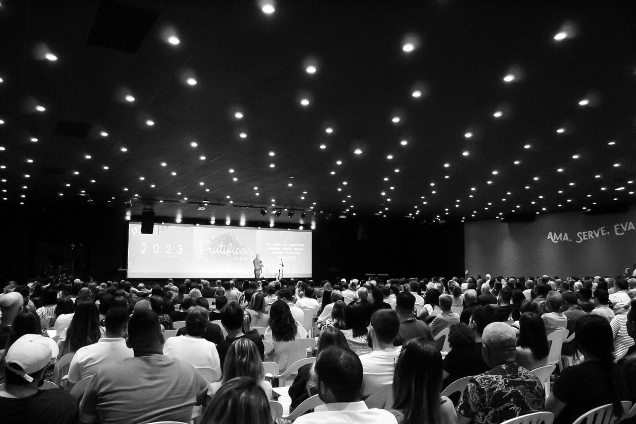 Uma imagem da igreja, tirada do fundo, mostrando a igreja cheia, com as luzes apagadas, e o povo de mãos dadas, com o Pastor Junior falando ao micronone na frente.